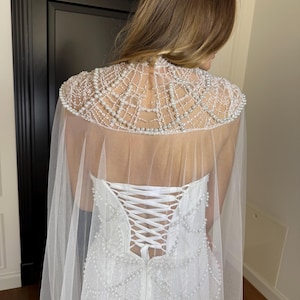 Strapless pearl lace bridal gown with sculpted corset, elegant mermaid silhouette and flowing sheer cape for contemporary weddings.