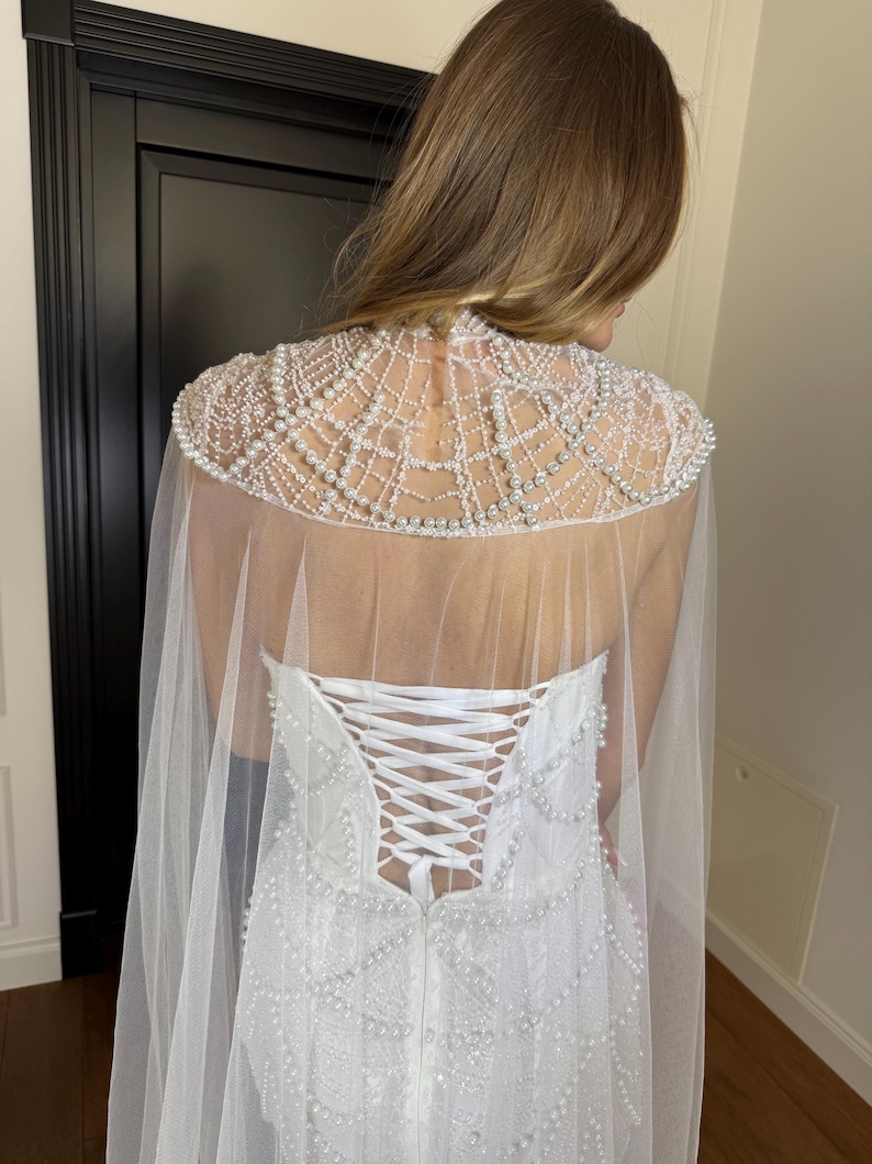 Strapless pearl lace bridal gown with sculpted corset, elegant mermaid silhouette and flowing sheer cape for contemporary weddings.