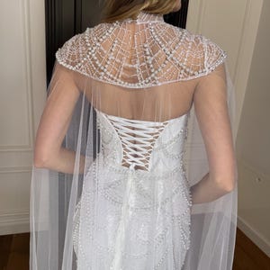 Couture-inspired lace wedding dress with fitted mermaid shape, pearl details and detachable sheer cape for statement bridal style.