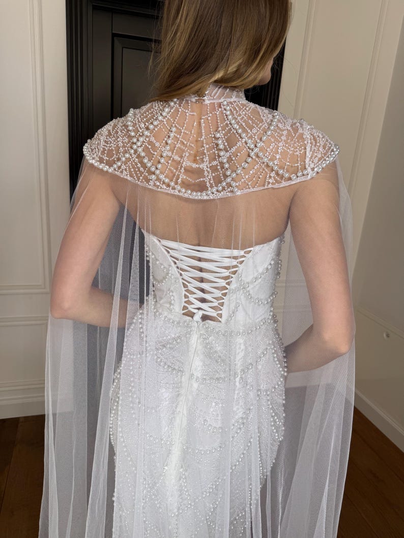 Couture-inspired lace wedding dress with fitted mermaid shape, pearl details and detachable sheer cape for statement bridal style.