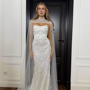 Pearl mermaid bridal gown featuring a structured corset, fitted silhouette and dramatic high-neck tulle cape with flowing panels.