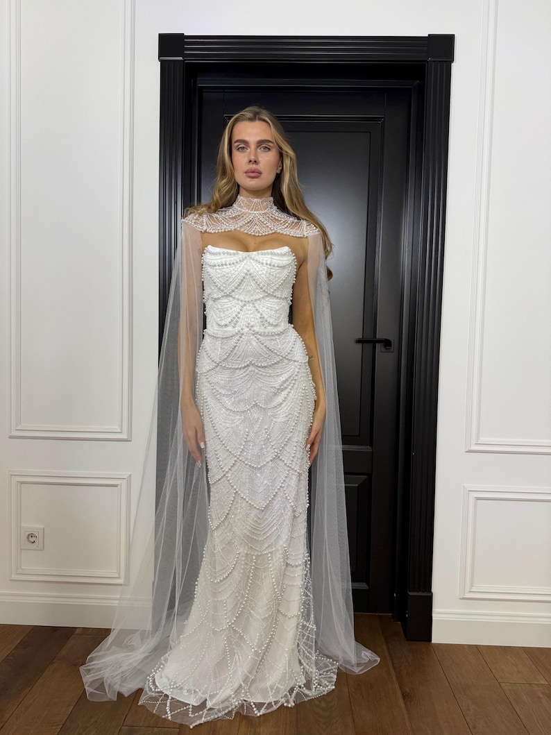 Pearl mermaid bridal gown featuring a structured corset, fitted silhouette and dramatic high-neck tulle cape with flowing panels.