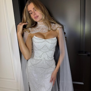 Elegant mermaid wedding dress with pearl detailing, lace embroidery and couture-inspired sheer cape for modern brides.