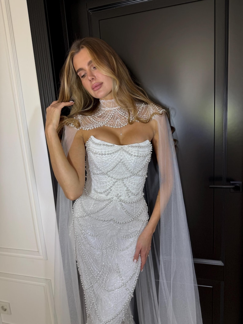 Elegant mermaid wedding dress with pearl detailing, lace embroidery and couture-inspired sheer cape for modern brides.