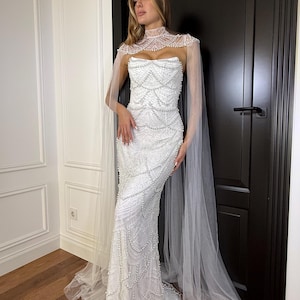 Fitted lace bridal gown with sculpted corset, pearl embellishments and flowing tulle cape for outdoor or garden weddings.