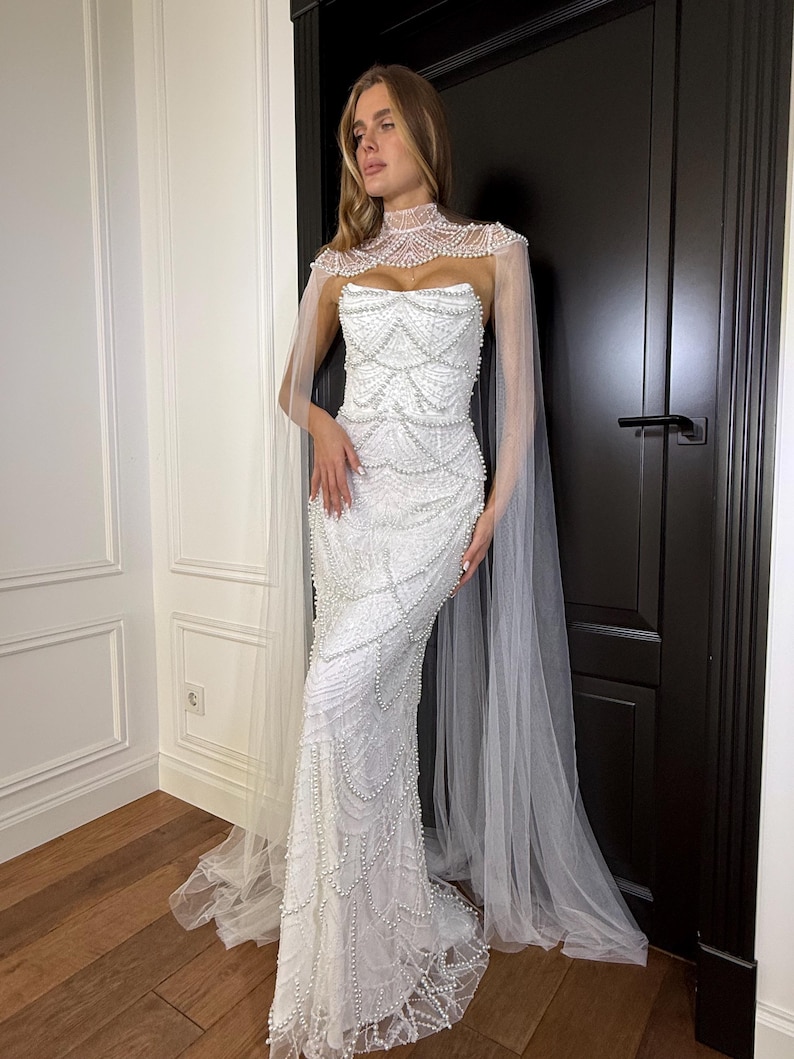 Fitted lace bridal gown with sculpted corset, pearl embellishments and flowing tulle cape for outdoor or garden weddings.