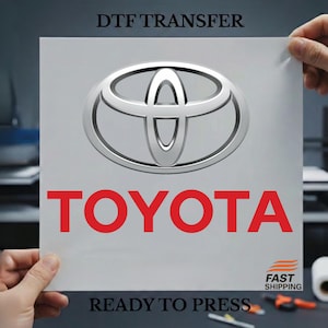 May include: A white DTF transfer sheet featuring the Toyota logo in silver and the word "TOYOTA" in red. The text "READY TO PRESS" is at the bottom. The sheet is held by two hands.