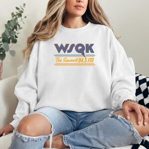 May include: White sweatshirt with the text "WSQK The Squawk 94.5 FM" in blue, orange, and teal. The sweatshirt is long-sleeved and has a crew neck. The person in the image is wearing blue jeans.