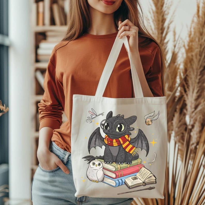 May include: Cream-colored tote bag featuring a cartoon dragon with a scarf, sitting on books. The dragon has large eyes and wings. An owl, a golden snitch, and a dragonfly are also depicted. The bag has white handles.