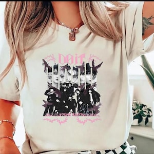 Pu&ograve; includere: T-shirt color crema con un design grafico. Il design include la parola "Dait" in rosa, immagini di volti e un gruppo di persone vestite di nero. &Egrave; visibile anche il testo "SKZ IT FAR STRAY KIDS".