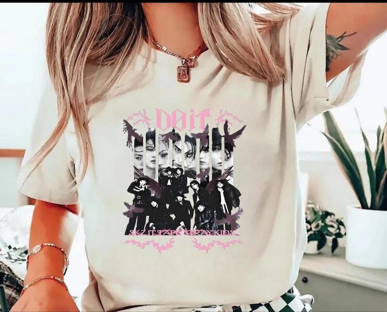 Pu&ograve; includere: T-shirt color crema con un design grafico. Il design include la parola "Dait" in rosa, immagini di volti e un gruppo di persone vestite di nero. &Egrave; visibile anche il testo "SKZ IT FAR STRAY KIDS".