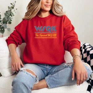 May include: A red sweatshirt with the text "WSQK The Squawk 94.5 FM" in blue, orange, and yellow. The sweatshirt is worn with blue jeans. The overall theme is radio station apparel.