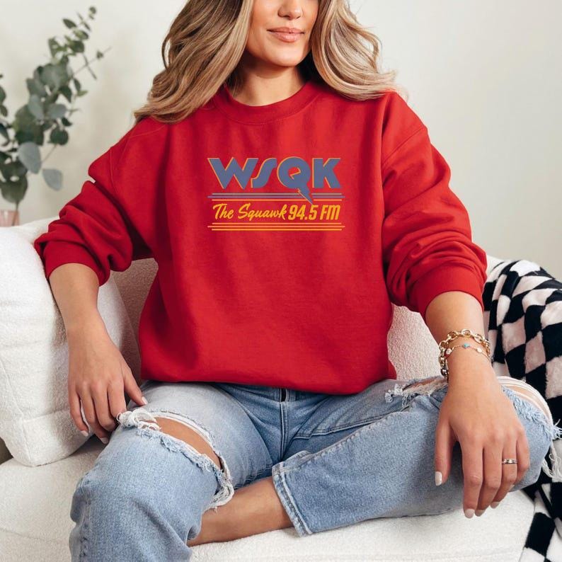 May include: A red sweatshirt with the text "WSQK The Squawk 94.5 FM" in blue, orange, and yellow. The sweatshirt is worn with blue jeans. The overall theme is radio station apparel.