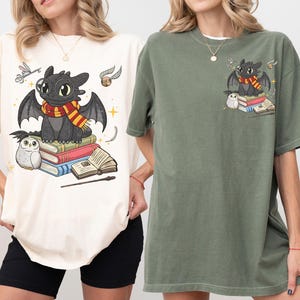 May include: Ivory and moss green t-shirts featuring a cartoon dragon with a scarf, sitting on books. The dragon is surrounded by a small owl, a wand, and other magical elements. The shirts are oversized and comfortable.