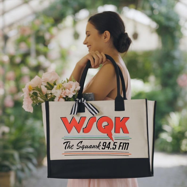 May include: A cream-colored tote bag with black handles and a black bottom panel. The bag features the text "WSQK The Squawk 94.5 FM" in red, black, and white. A bouquet of pink flowers and a black and white striped bow are visible.
