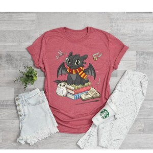 May include: Red t-shirt featuring a cartoon dragon with a scarf, sitting on books. The dragon is black with large eyes and wings. An owl, a wand, and other graphics are also on the shirt. The shirt is paired with denim shorts and a white sweater.