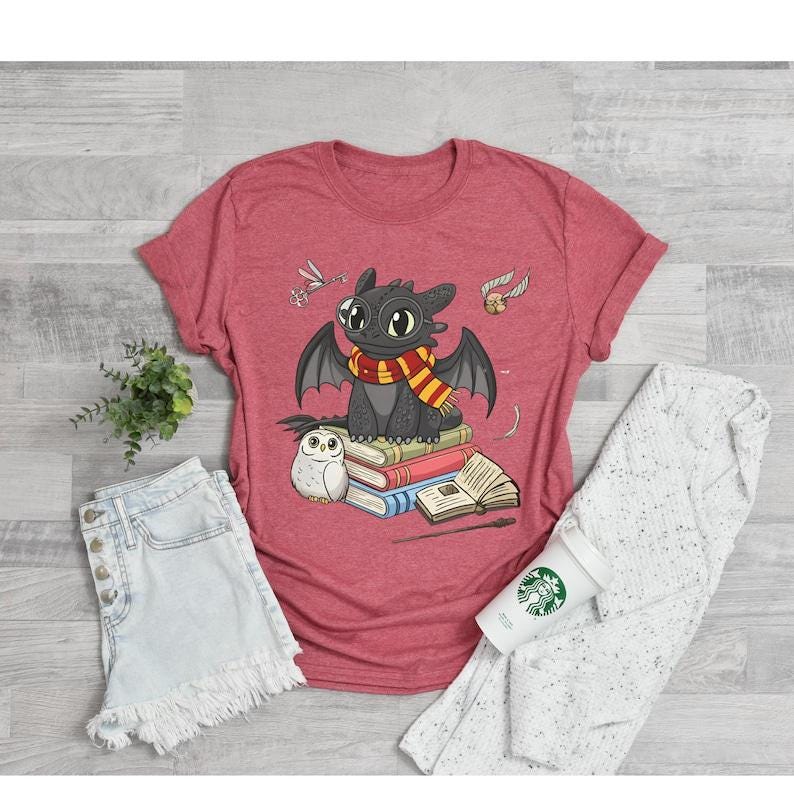 May include: Red t-shirt featuring a cartoon dragon with a scarf, sitting on books. The dragon is black with large eyes and wings. An owl, a wand, and other graphics are also on the shirt. The shirt is paired with denim shorts and a white sweater.