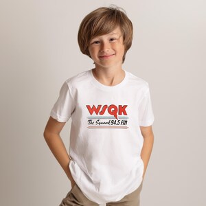 May include: A white t-shirt featuring the text "WSQK The Squawk 94.5 FM" in red and blue. The shirt has a crew neck and short sleeves. The design includes a red stylized "Q" and two horizontal lines.