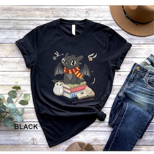 May include: Black t-shirt featuring a cartoon dragon wearing a scarf, sitting on books, with an owl and other graphics. The word "BLACK" is printed at the bottom left of the shirt. The shirt is folded on a wooden surface.