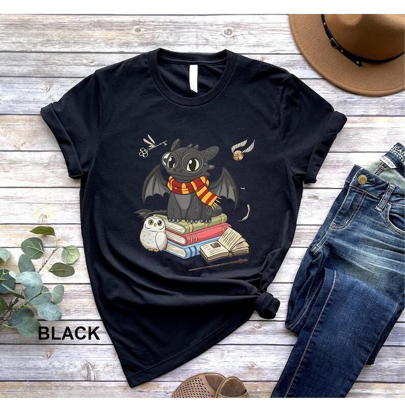 May include: Black t-shirt featuring a cartoon dragon wearing a scarf, sitting on books, with an owl and other graphics. The word "BLACK" is printed at the bottom left of the shirt. The shirt is folded on a wooden surface.