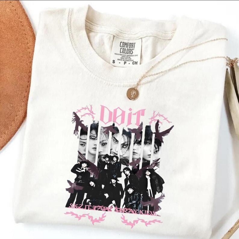 Pu&ograve; includere: T-shirt color crema con una grafica raffigurante un gruppo di persone e la parola "Doit" in rosa. Il testo "SKZ ITTARAE STRAY KIDS" &egrave; in basso. Una collana d'oro con un ciondolo &egrave; drappeggiata sulla maglietta.