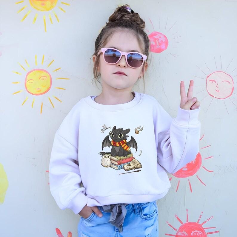 May include: A white sweatshirt featuring a cartoon dragon with a red and yellow scarf, perched on books. The dragon has black wings and is next to an owl. The background has drawn suns and pink sunglasses.