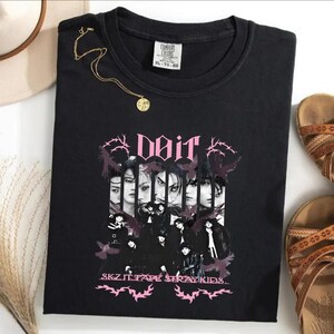 Pu&ograve; includere: T-shirt nero girocollo con grafica rosa "Doit", collage di volti e testo "SKZ IT TAPE STRAY KIDS". Visibili anche una collana d'oro e sandali.