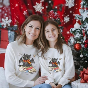 May include: Two people wearing matching white sweatshirts with a cartoon dragon design. The dragon has black wings, a red and gold scarf, and is sitting on books. An owl and other magical elements are also featured. The background includes a Christmas tree and red decorations.
