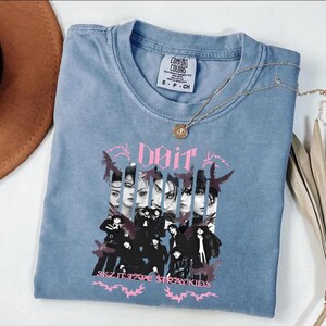 Pu&ograve; includere: T-shirt blu a maniche corte con un design grafico raffigurante un gruppo di persone e la parola "Doit" in rosa. Il testo "SKZ IT TAPE STRAY KIDS" &egrave; stampato anche sulla maglietta. Una collana d'oro &egrave; drappeggiata sulla maglietta.