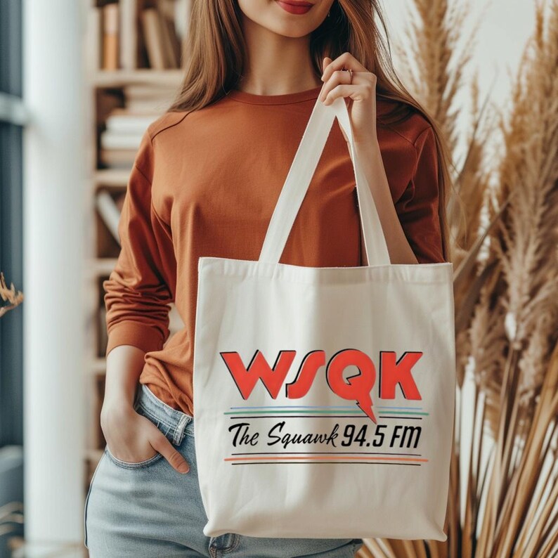 May include: Cream-colored tote bag with the text "WSQK The Squawk 94.5 FM" in red, black, and green. The bag has white handles and is held by a person wearing a rust-colored long-sleeve shirt and blue jeans.