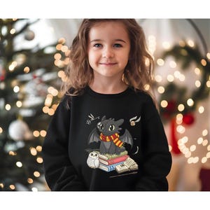 May include: A black sweatshirt featuring a cartoon dragon wearing a scarf, sitting on books with an owl. The design includes a golden snitch and other magical elements. The sweatshirt is worn by a young girl.