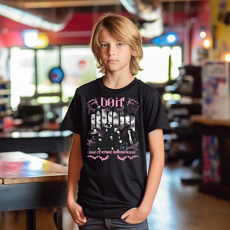 Pu&ograve; includere: T-shirt nera con un design grafico rosa. Il design include la parola "Doir", immagini di volti e il testo "SKZ ITZY STRAYKIDS". La t-shirt &egrave; indossata da una persona.