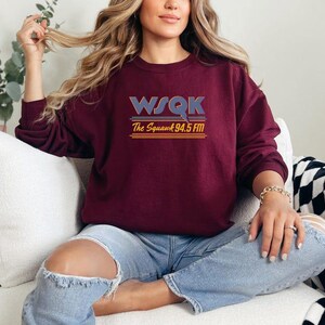 May include: A maroon sweatshirt with the text "WSQK The Squawk 94.5 FM" in a retro font. The sweatshirt has long sleeves and a crew neck. The person is wearing blue jeans.
