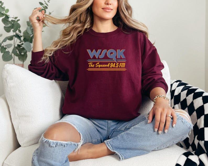 May include: A maroon sweatshirt with the text "WSQK The Squawk 94.5 FM" in a retro font. The sweatshirt has long sleeves and a crew neck. The person is wearing blue jeans.
