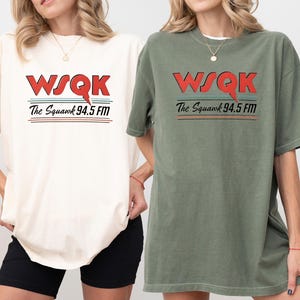 May include: Two short-sleeved t-shirts, one ivory and one moss green, each featuring the text "WSQK The Squawk 94.5 FM" in red, black, and teal. The shirts are a relaxed fit.
