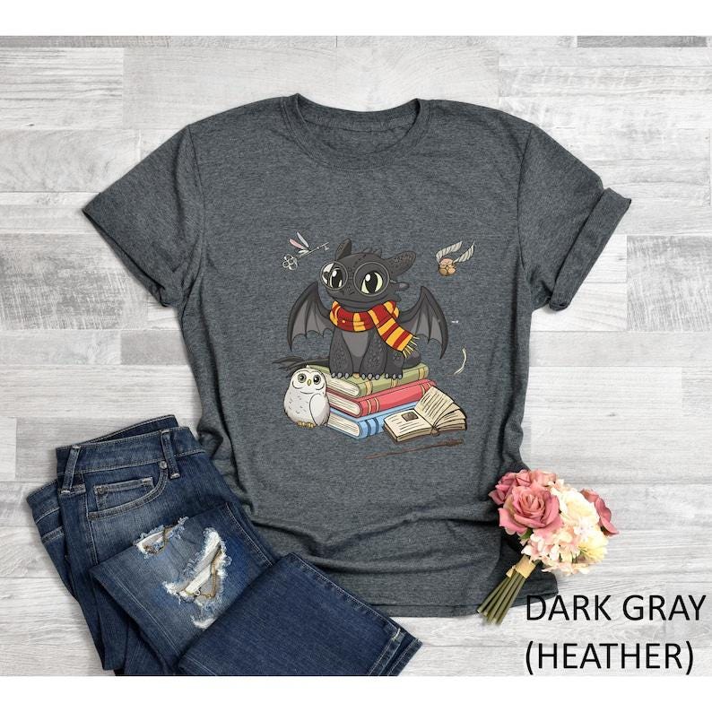 May include: Dark gray heather t-shirt featuring a cartoon dragon wearing a scarf, sitting on books, with an owl and other graphics. The text "DARK GRAY (HEATHER)" is printed on the bottom right of the image. The shirt is paired with blue jeans.