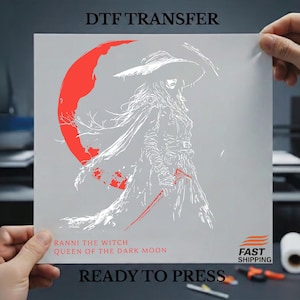 May include: A DTF transfer featuring a white illustration of a witch with a large hat, cape, and sword, set against a gray background with a red crescent moon. Text includes "RANNI THE WITCH QUEEN OF THE DARK MOON" and "READY TO PRESS".