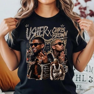 Pode incluir: Uma t-shirt preta com uma estampa gr&aacute;fica de Usher e Chris Brown. O design inclui detalhes de correntes douradas, coroas e o texto "USHER X CHRIS BROWN" juntamente com t&iacute;tulos de m&uacute;sicas como "My Way" e "Confessions" em dourado.