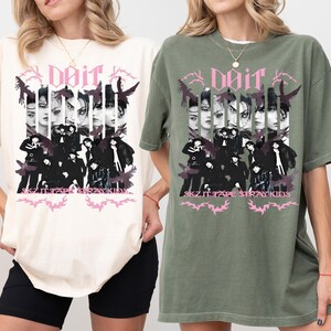 Pu&ograve; includere: T-shirt avorio e verde muschio con un design grafico. Il design presenta il testo "doir" e "SKZ IT TAPE STRAY KIDS", insieme a immagini di persone ed elementi decorativi in nero e rosa.