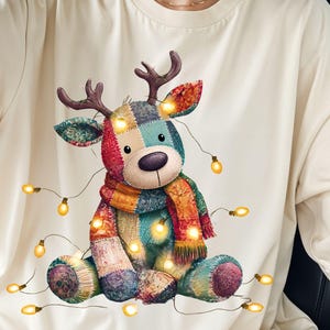 May include: Cream-colored t-shirt featuring a colorful patchwork reindeer design with a scarf and string lights. The reindeer has brown antlers and is surrounded by glowing yellow lights, creating a festive holiday theme.