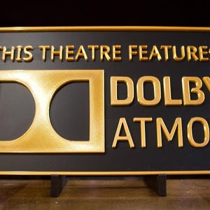 Op de afbeelding: Een rechthoekig zwart bord met gouden letters en rand. De tekst luidt "THIS THEATRE FEATURES DOLBY ATMOS" met het Dolby-logo. Het bord is gemonteerd op een kleine zwarte standaard.