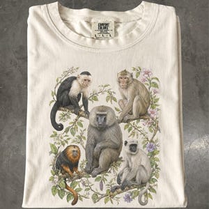 Op de afbeelding: Een crèmekleurig t-shirt met een Comfort Colors label in maat L. De afbeelding toont vijf verschillende apen die tussen groene bladeren en roze en paarse bloemen zitten. De apen zijn gedetailleerd afgebeeld met verschillende vachtkleuren.