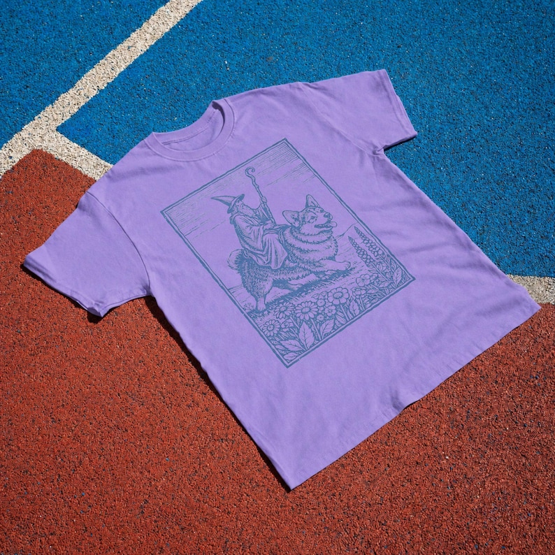 May include: A light purple short-sleeve t-shirt featuring a dark purple rectangular graphic print. The illustration depicts a figure wearing a pointed hat and robe riding a corgi dog, surrounded by a field of flowers within a thin border.