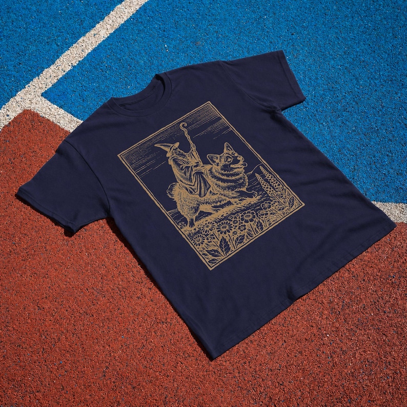 May include: A navy blue short-sleeved t-shirt featuring a gold-colored graphic print of a wizard riding a corgi dog. The illustration is framed within a rectangular border and depicts the figures standing in a field of flowers and foliage.