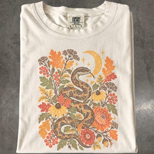 以下が含まれることがあります： クリーム色のTシャツで、とぐろを巻いたヘビと、オレンジや黄色の花、オークの葉、ベリー、どんぐり、三日月といった秋をテーマにした要素が描かれたグラフィックプリントが施されています。シャツにはComfort Colorsのブランドラベルが付いています。