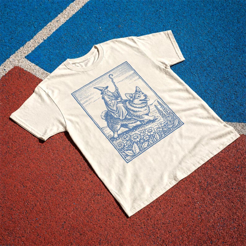May include: A cream-colored short-sleeve t-shirt featuring a blue graphic print of a wizard riding a corgi. The illustration is framed in a rectangular border and depicts the wizard holding a staff while sitting atop the dog in a field of flowers.