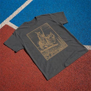 May include: A charcoal gray short-sleeve t-shirt featuring a gold-toned line art graphic. The illustration depicts a robed figure wearing a pointed hat and holding a staff, standing beside a corgi dog amidst a field of flowers and foliage.