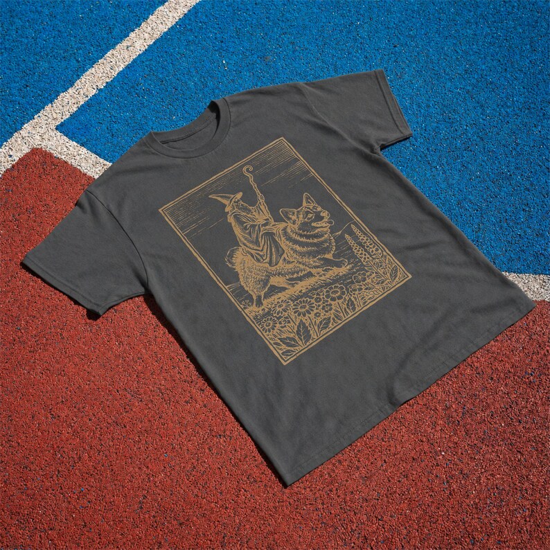 May include: A charcoal gray short-sleeve t-shirt featuring a gold-toned line art graphic. The illustration depicts a robed figure wearing a pointed hat and holding a staff, standing beside a corgi dog amidst a field of flowers and foliage.
