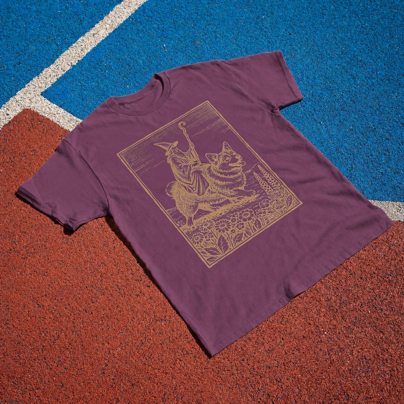 May include: A maroon short-sleeved t-shirt featuring a gold-colored line art graphic. The illustration depicts a robed figure wearing a pointed hat and holding a staff, standing beside a fluffy dog in a field of flowers under a lined sky.