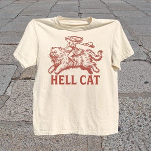 Könnte beinhalten: Cremefarbenes T-Shirt mit roter Grafik einer Person, die auf einer großen Katze reitet, darunter der Text "HELL CAT". Das Design ist eine Illustration im Vintage-Stil. Das Shirt hat kurze Ärmel und einen Rundhalsausschnitt.